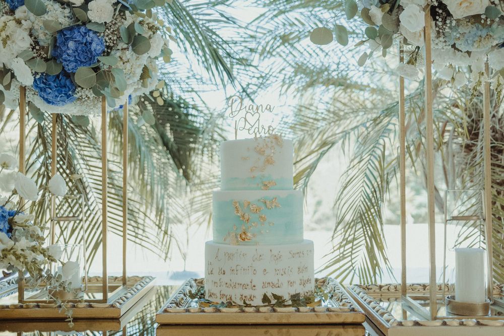 Elegant four-tier wedding cake with gold details displayed under tropical palm leaves and blue and white floral arrangements.