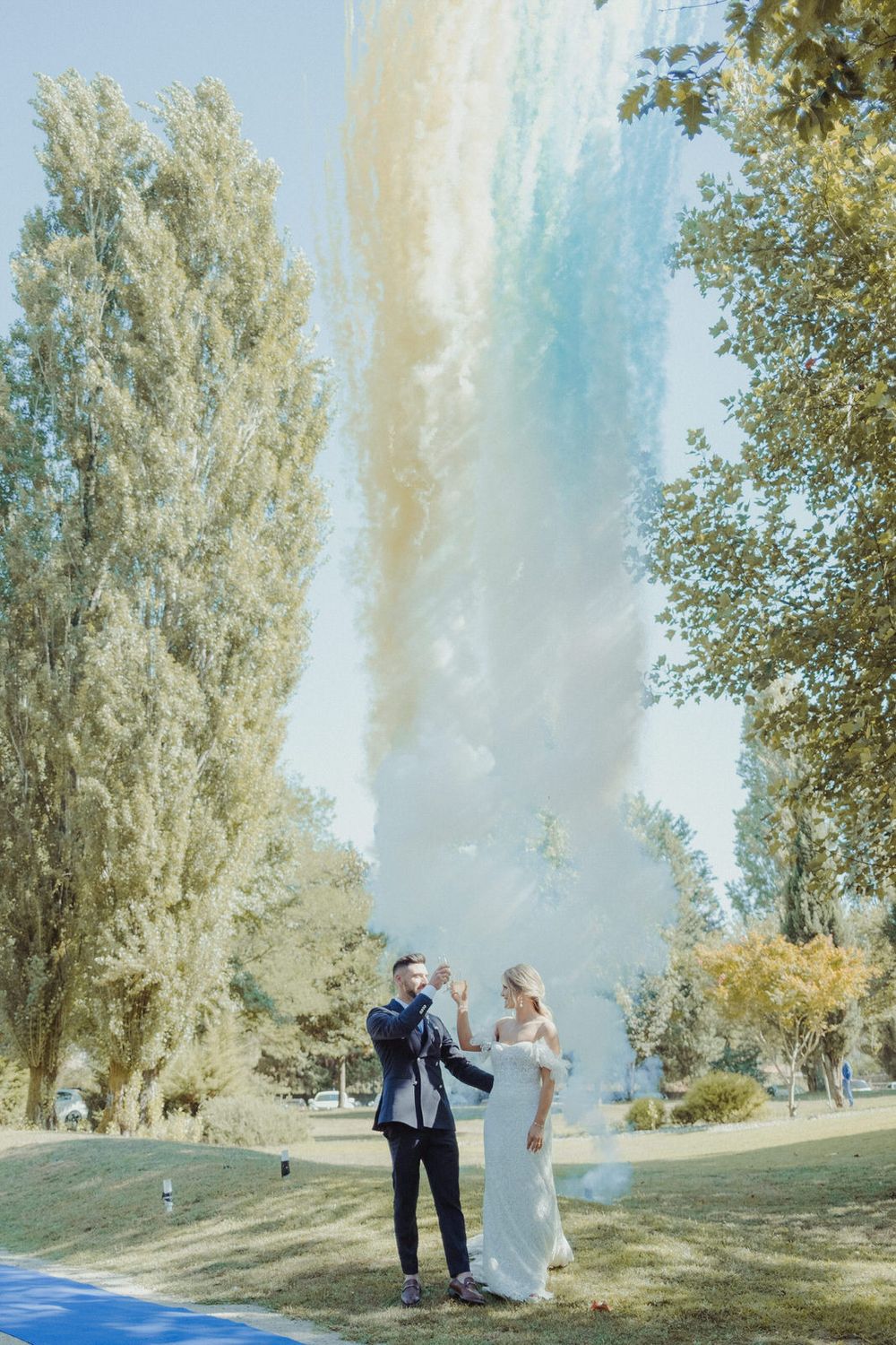 Bride and groom celebrate with colorful smoke bombs in a lush green park on their wedding day.