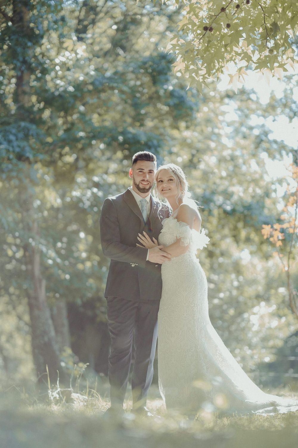 Bride in white gown and groom in gray suit embrace outdoors under sunlit trees in a romantic wedding portrait.