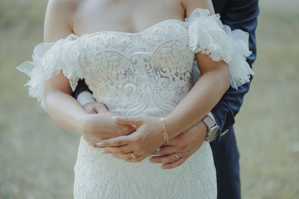 Bride in elegant off-shoulder lace wedding gown with groom embracing her from behind, showcasing intricate details.