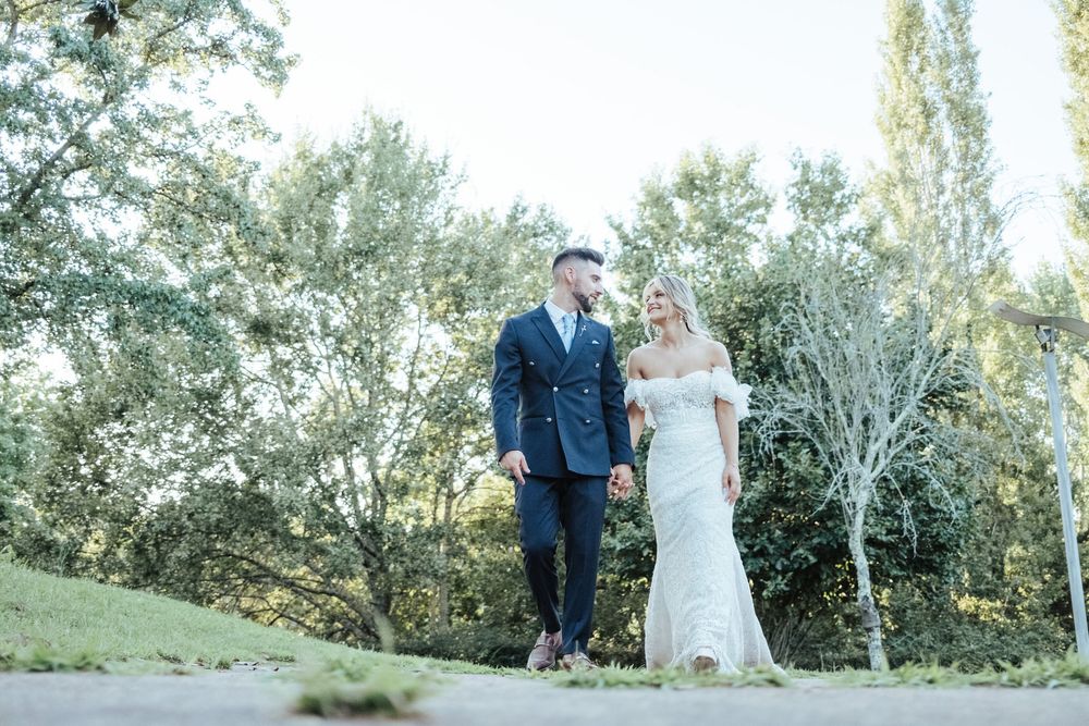 Bride and groom walk hand in hand through lush green park in elegant wedding attire.