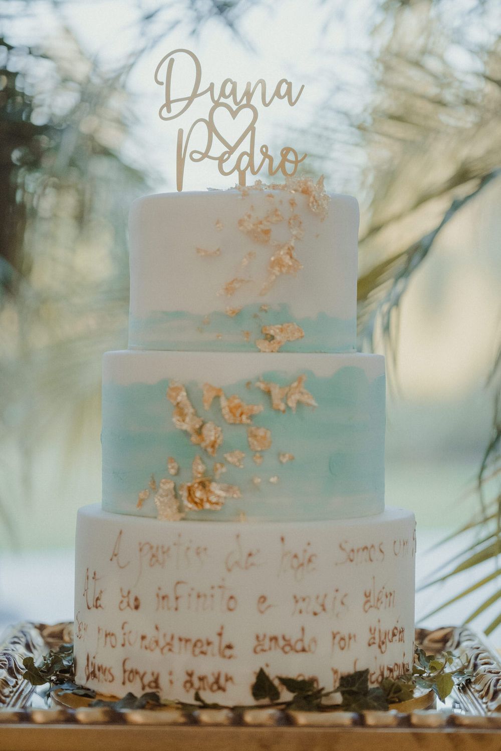 Elegant three-tier wedding cake with blue watercolor design, gold leaf accents, and Diana & Pedro cake topper.