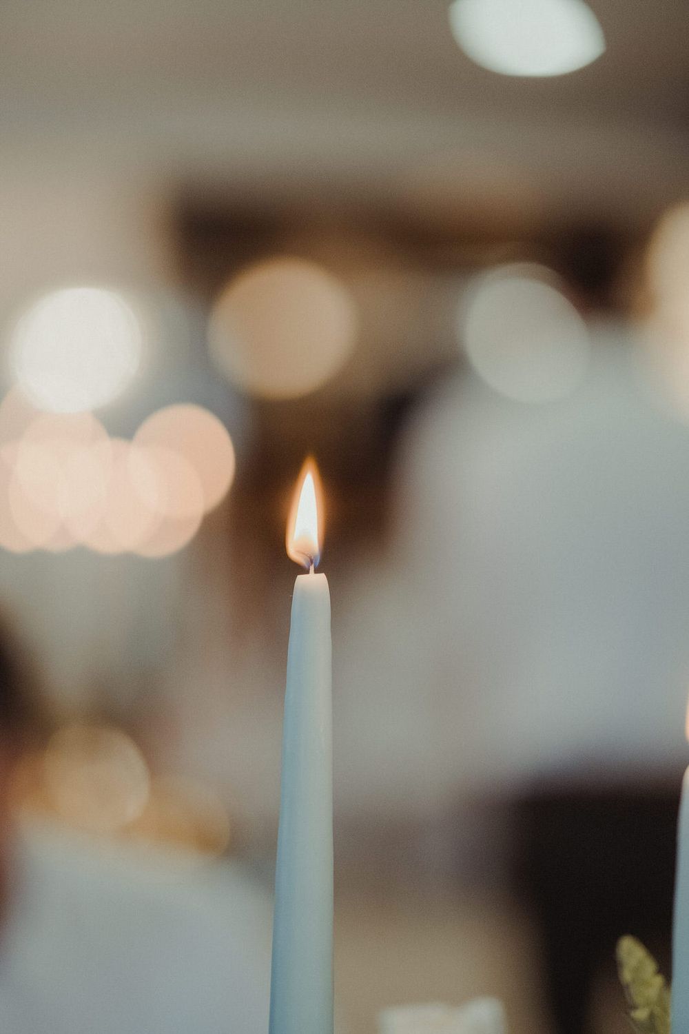 A single white taper candle with a glowing flame against a soft bokeh background of warm lights.