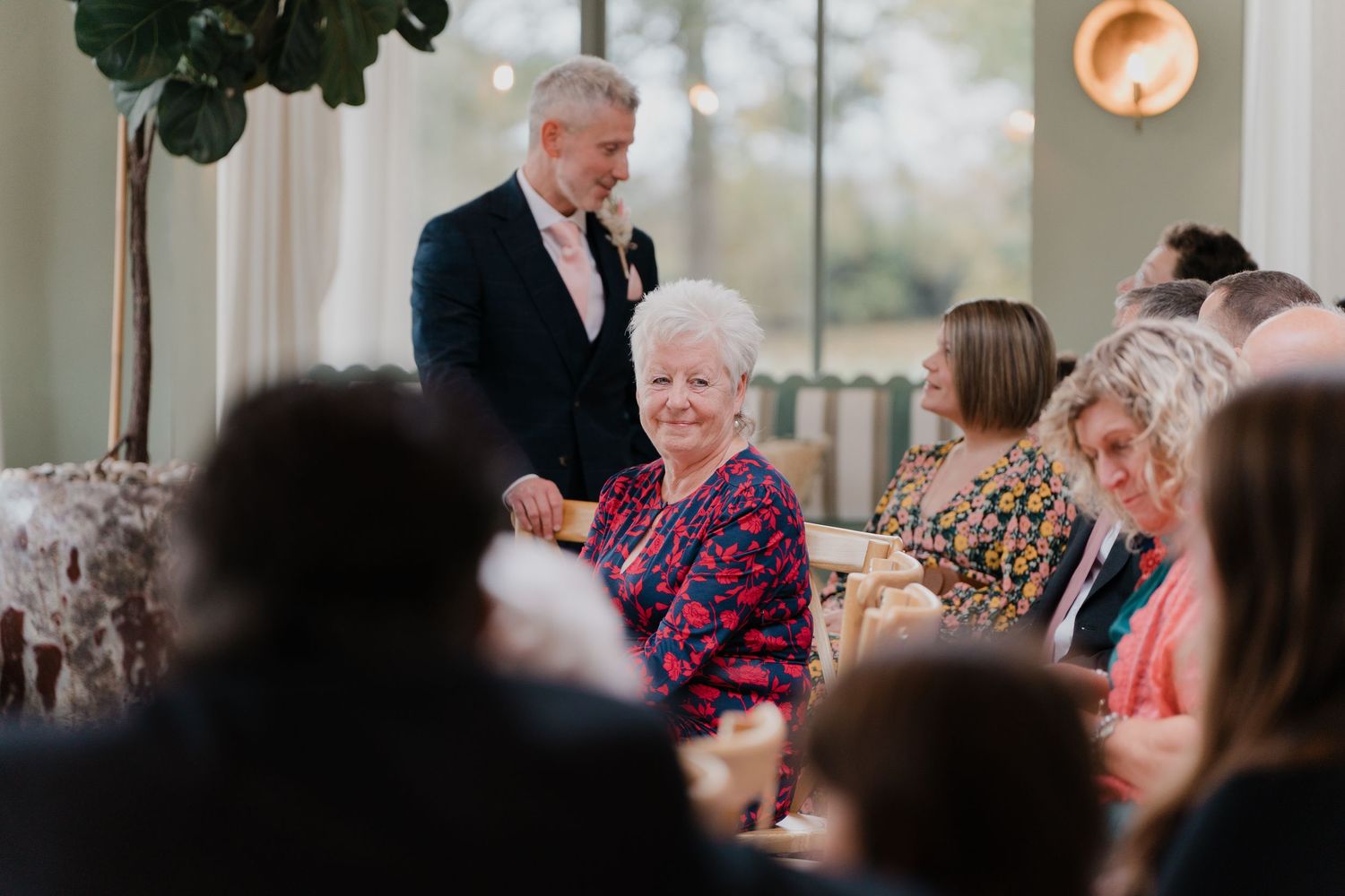 Elderly woman smiling at wedding reception seated among guests while groom stands nearby in navy suit.