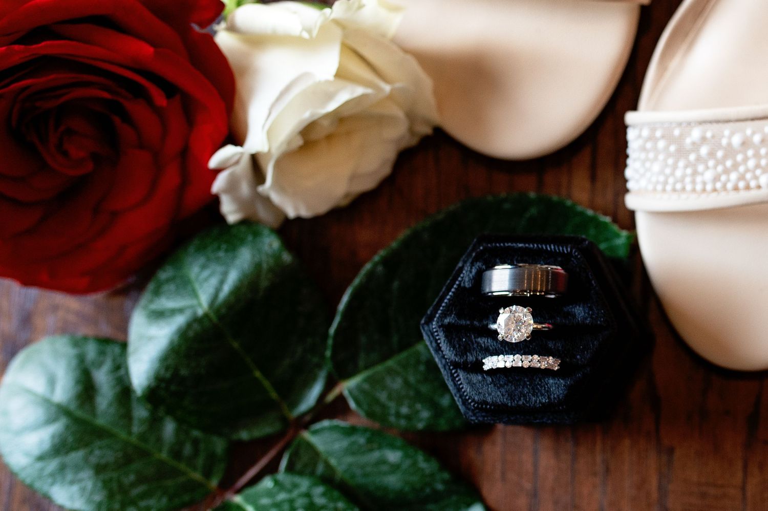Wedding rings in velvet box beside red and white roses and pearl bridal heels on wooden surface.