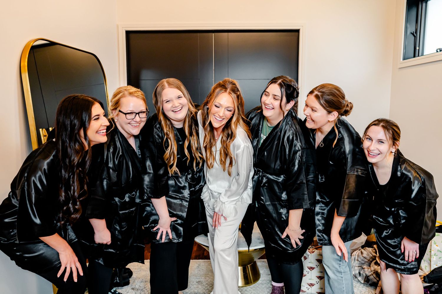 Bride in white robe smiling with six bridesmaids in black satin robes during wedding preparations.