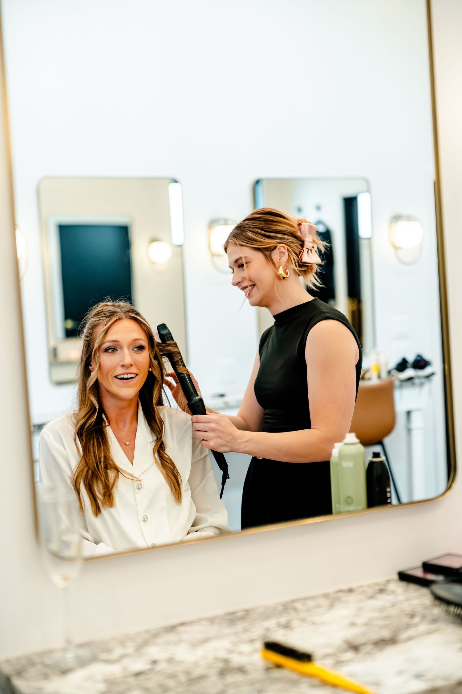 Smiling hairstylist curling bride's hair with a curling iron in a bright salon mirror reflection.