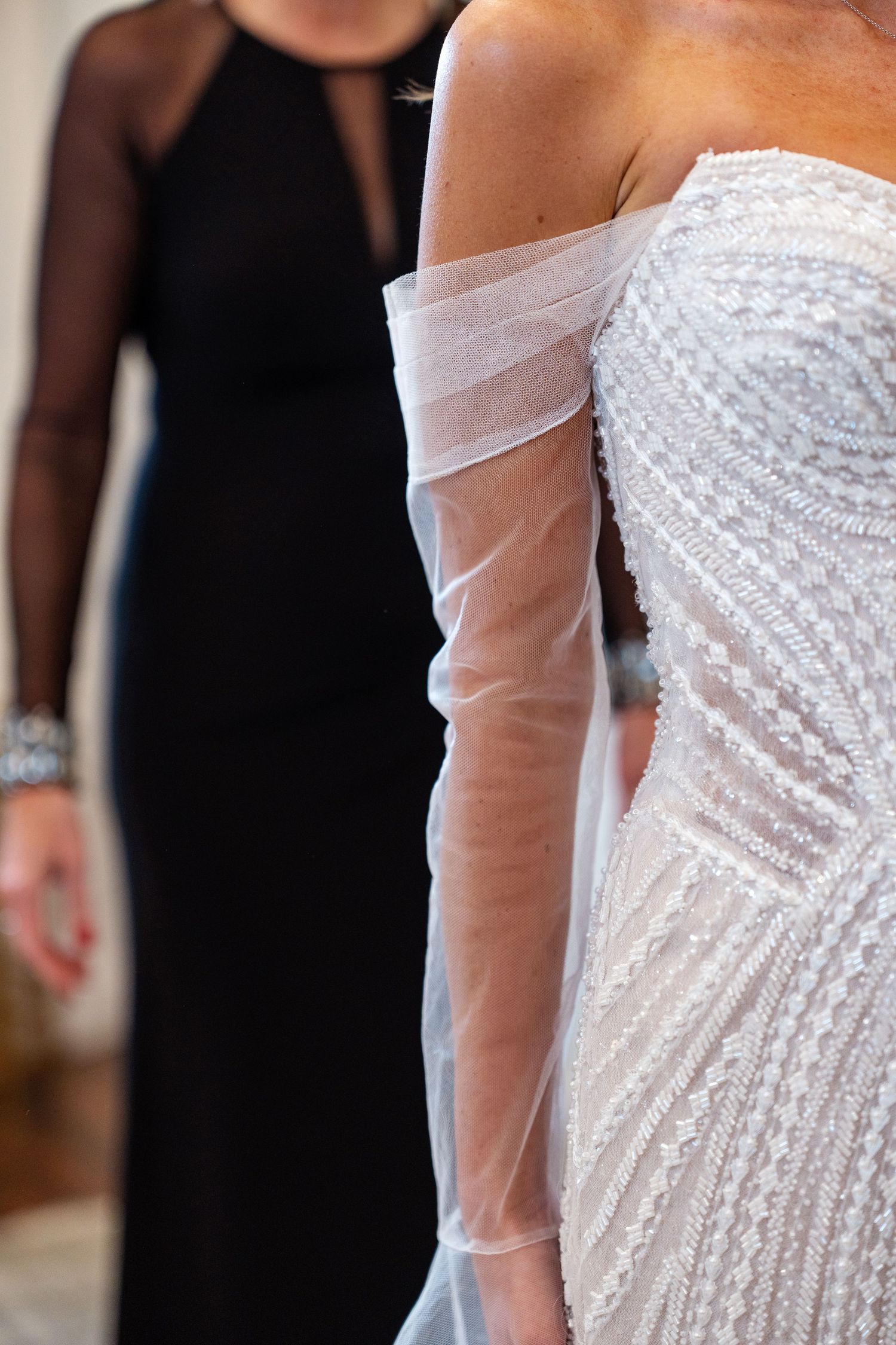 Close-up of a bride in a beaded off-shoulder white gown with sheer sleeves, beside a woman in a black dress.