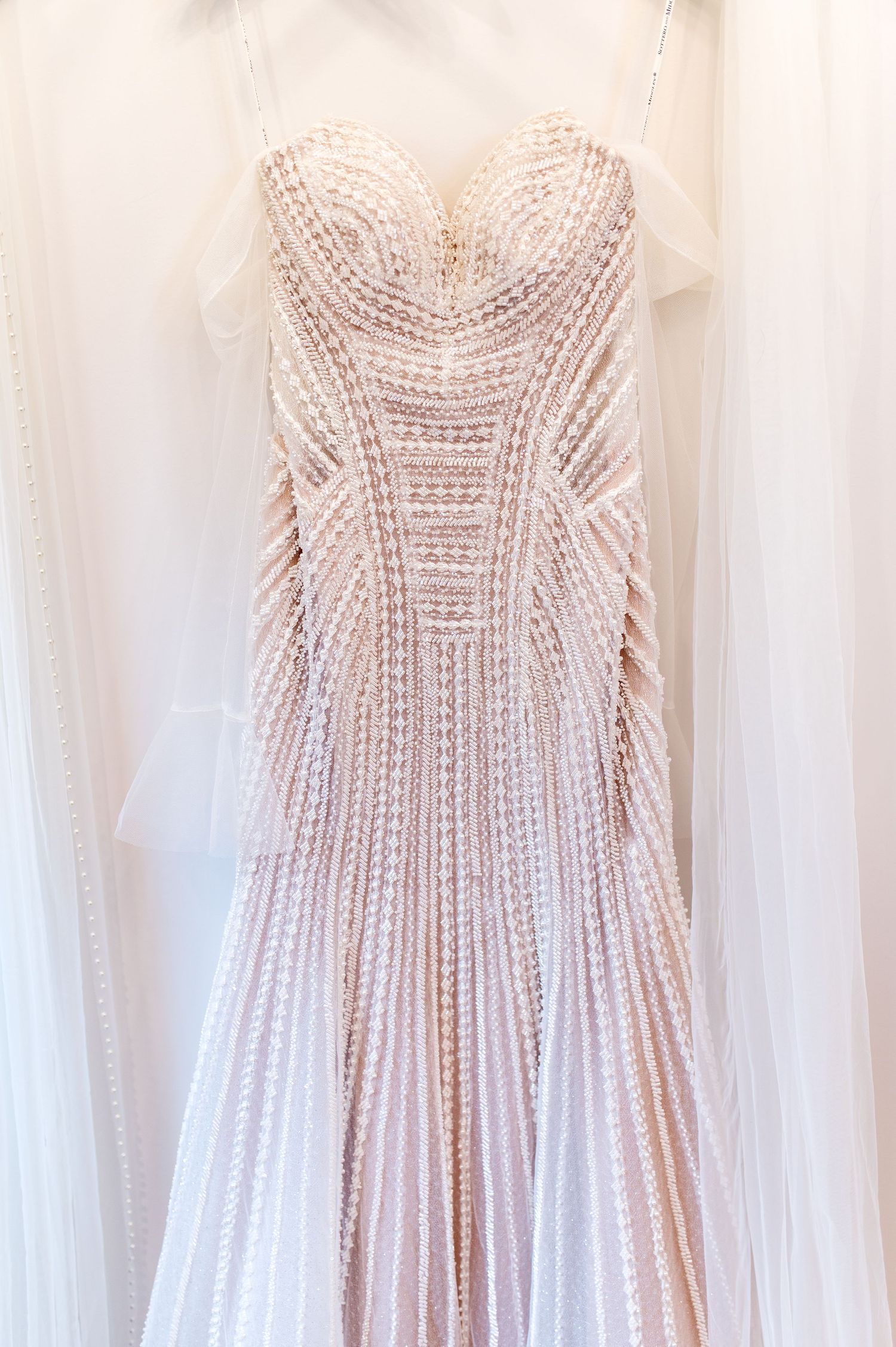 Elegant strapless beaded wedding gown with intricate pearl embroidery displayed on a hanger with sheer veil.