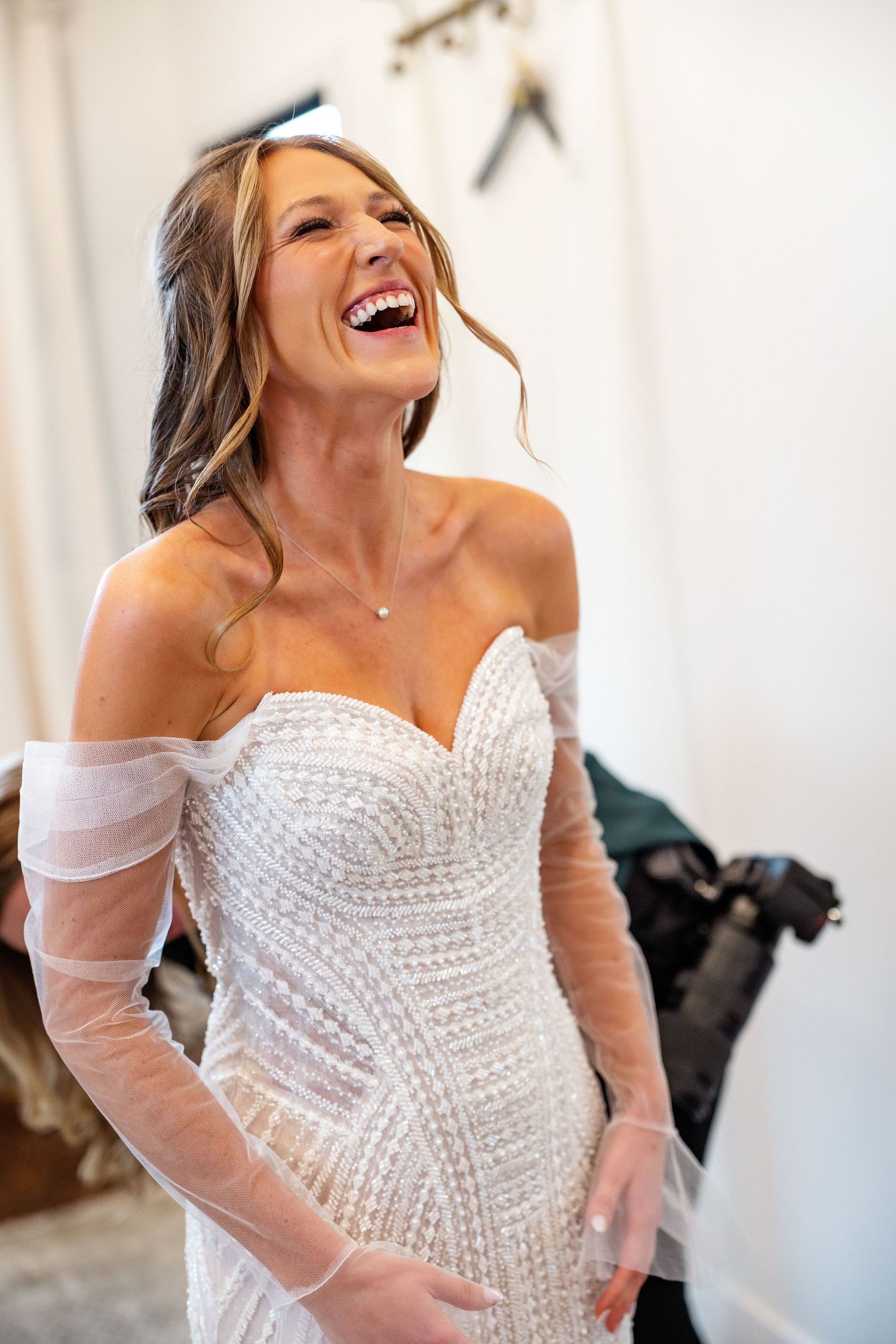 Radiant bride laughing joyfully in a beaded off-shoulder wedding gown with sheer sleeves during bridal prep.