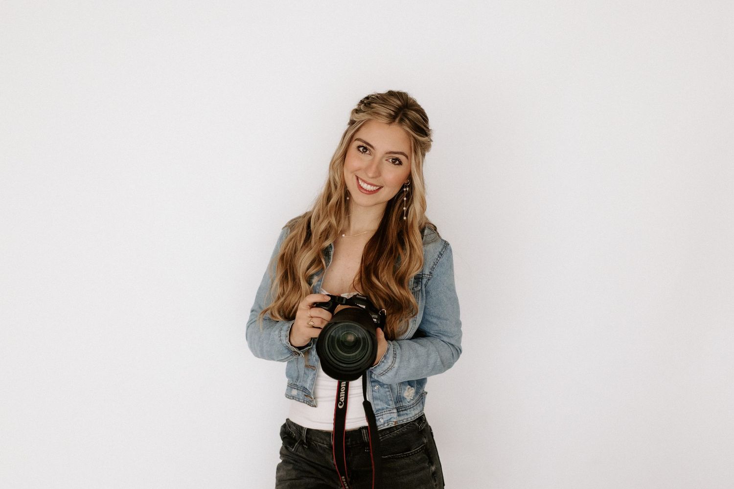 Smiling young woman with long wavy hair holding a DSLR camera, wearing a denim jacket, posed against a white background.