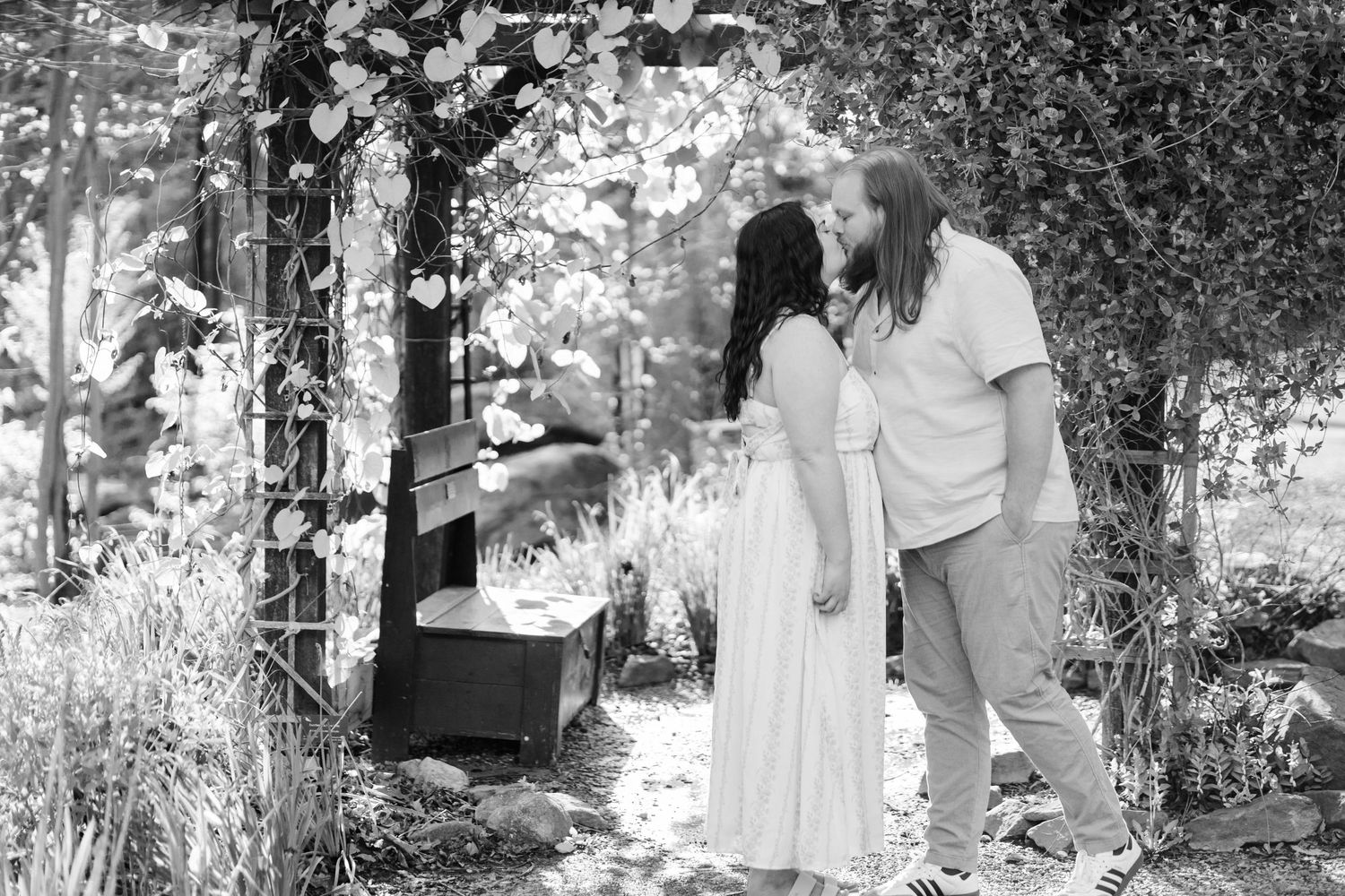 Black & white photo of a couple sharing a kiss under a garden arbor surrounded by lush greenery. morgantown wv botanical gardens engagement