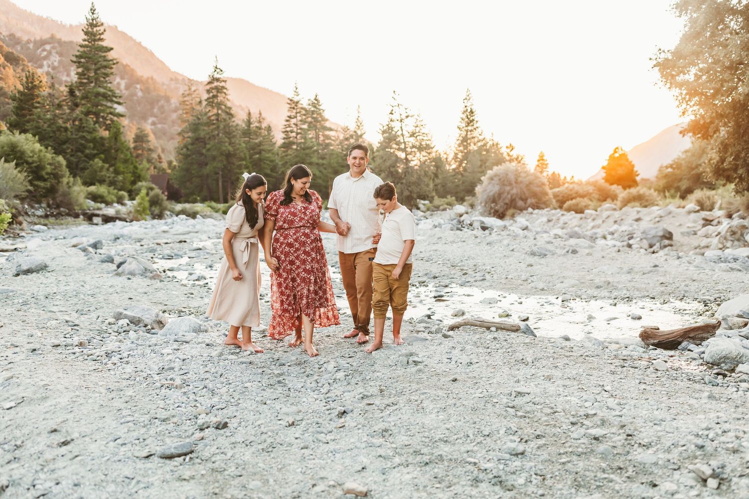 Family of four walking together by a rocky mountain river at golden hour sunset with pine trees.