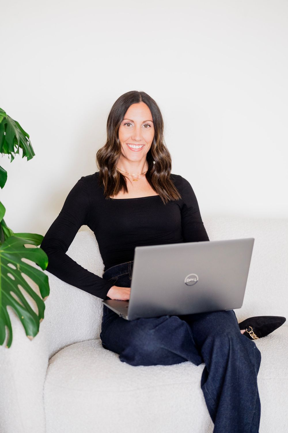 Professional brunette woman smiling with Dell laptop on white couch near monstera plant.