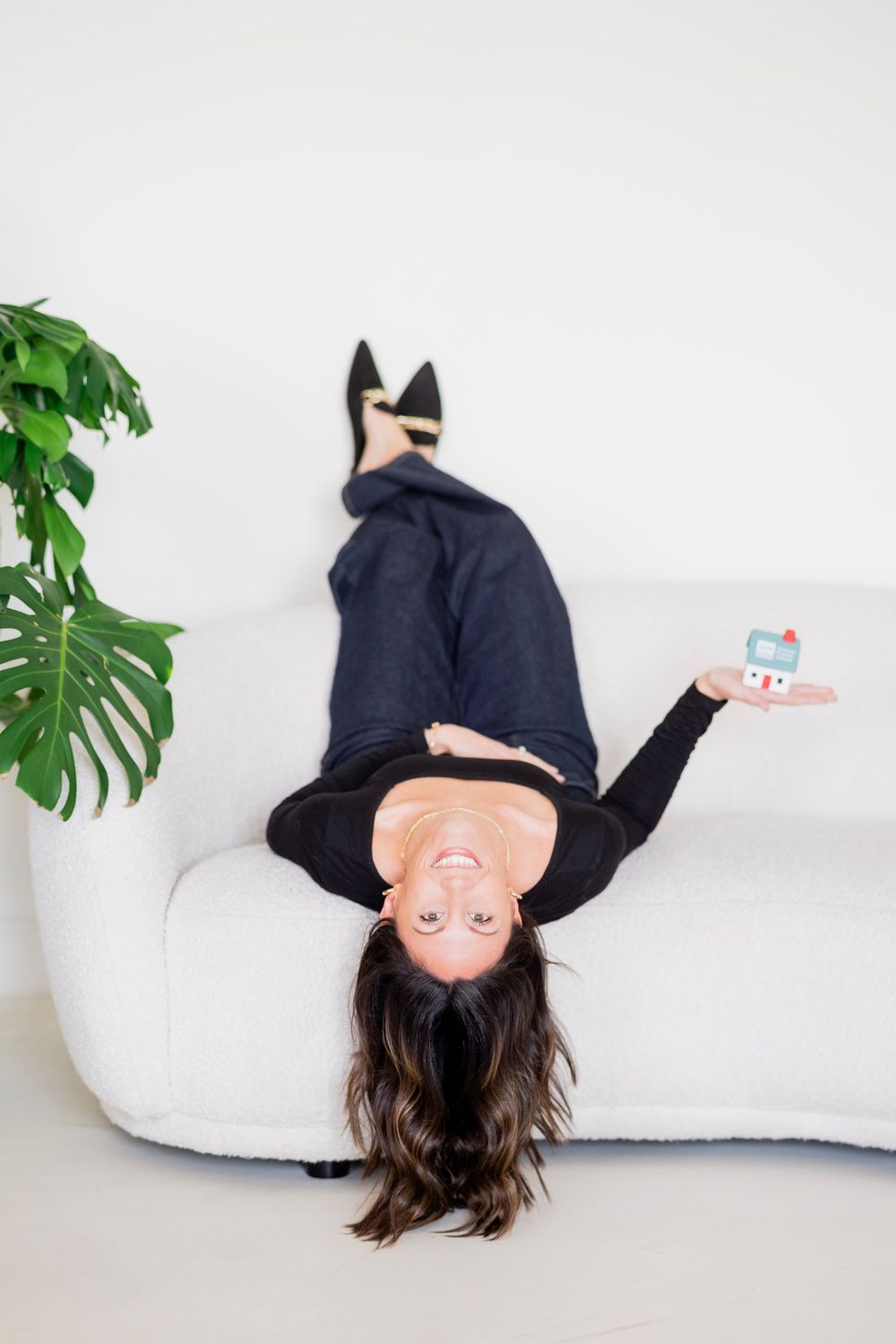 Woman lying upside down on white sofa holding a miniature house model, laughing playfully.