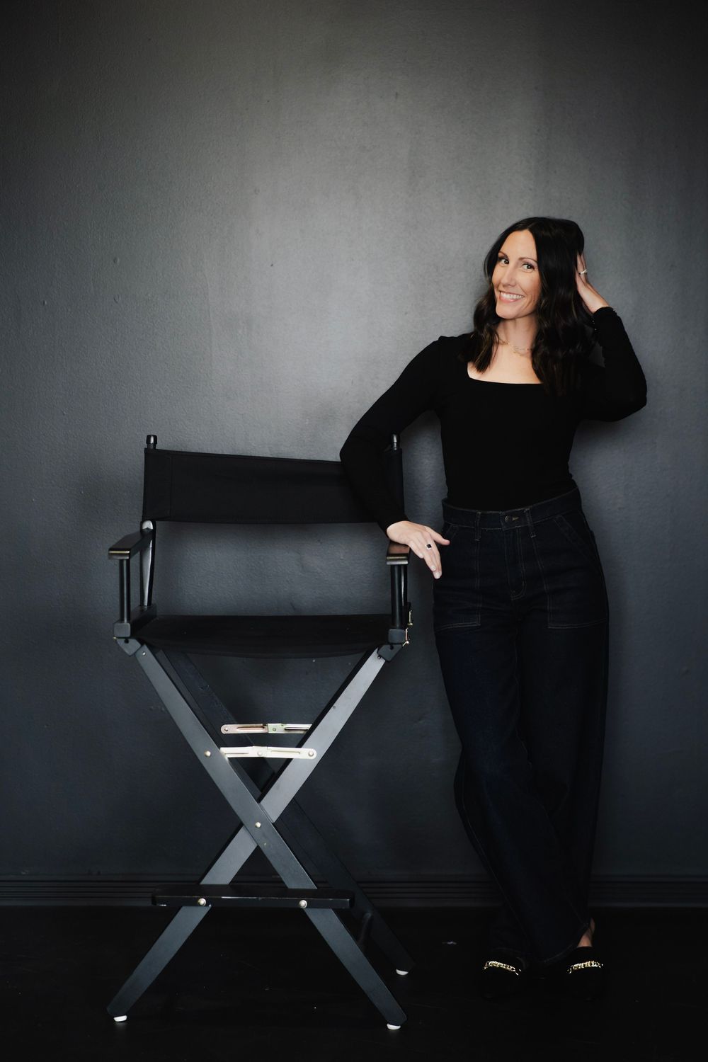 Stylish brunette woman in all-black outfit standing beside a director's chair against gray wall.