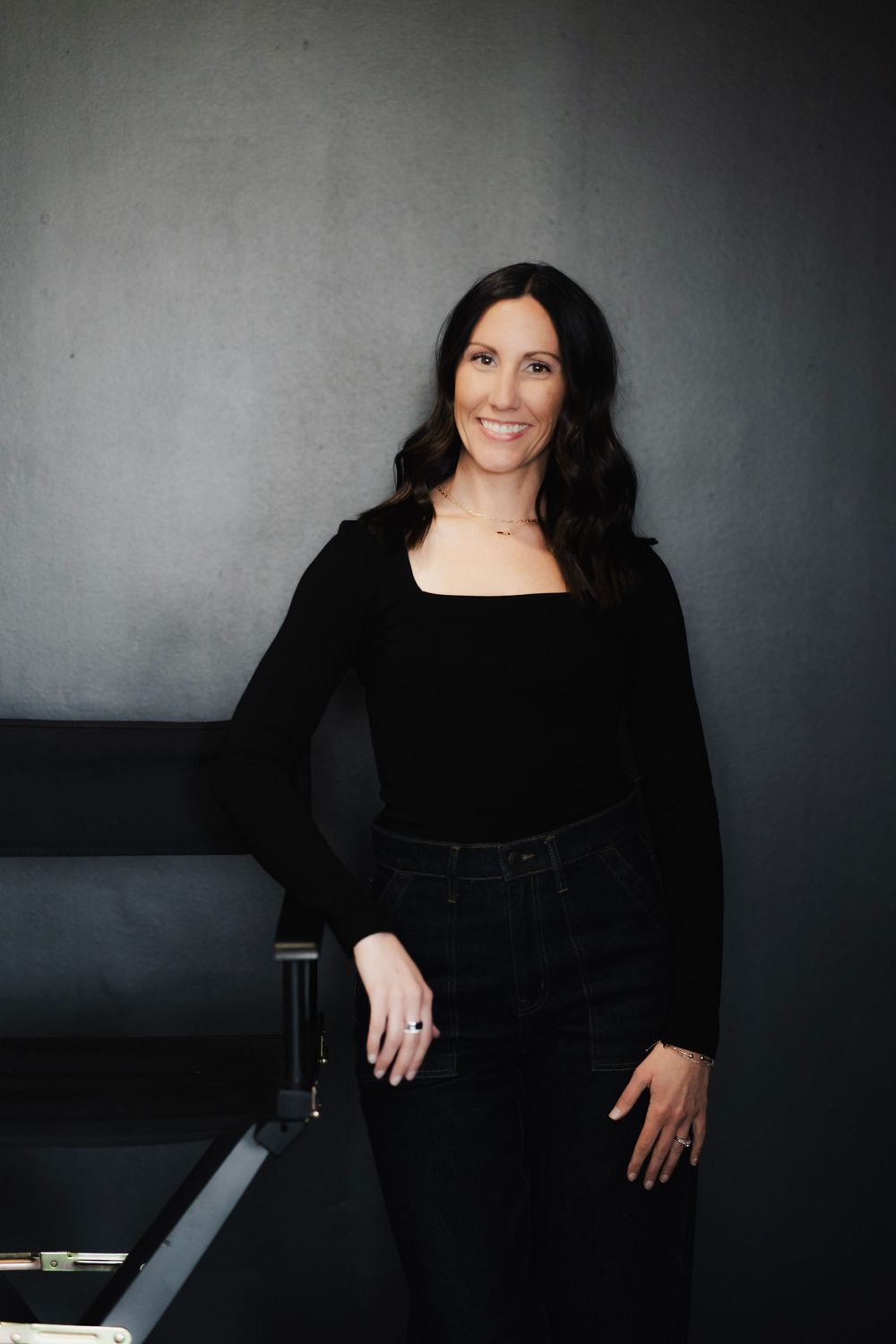 Professional brunette woman smiling in all-black outfit standing near director's chair on gray background.