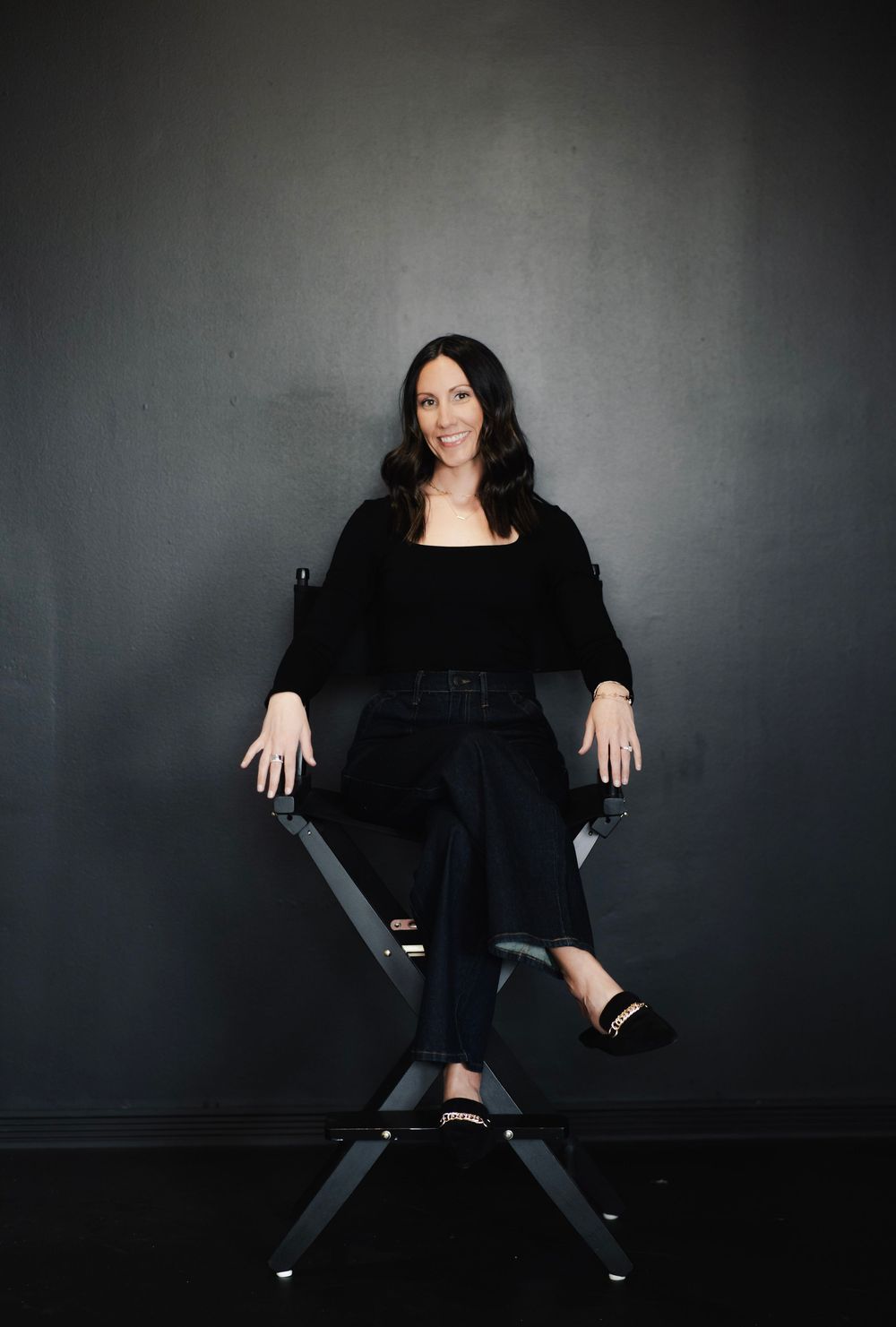 Smiling brunette woman sitting confidently in a director's chair against a dark gray background.