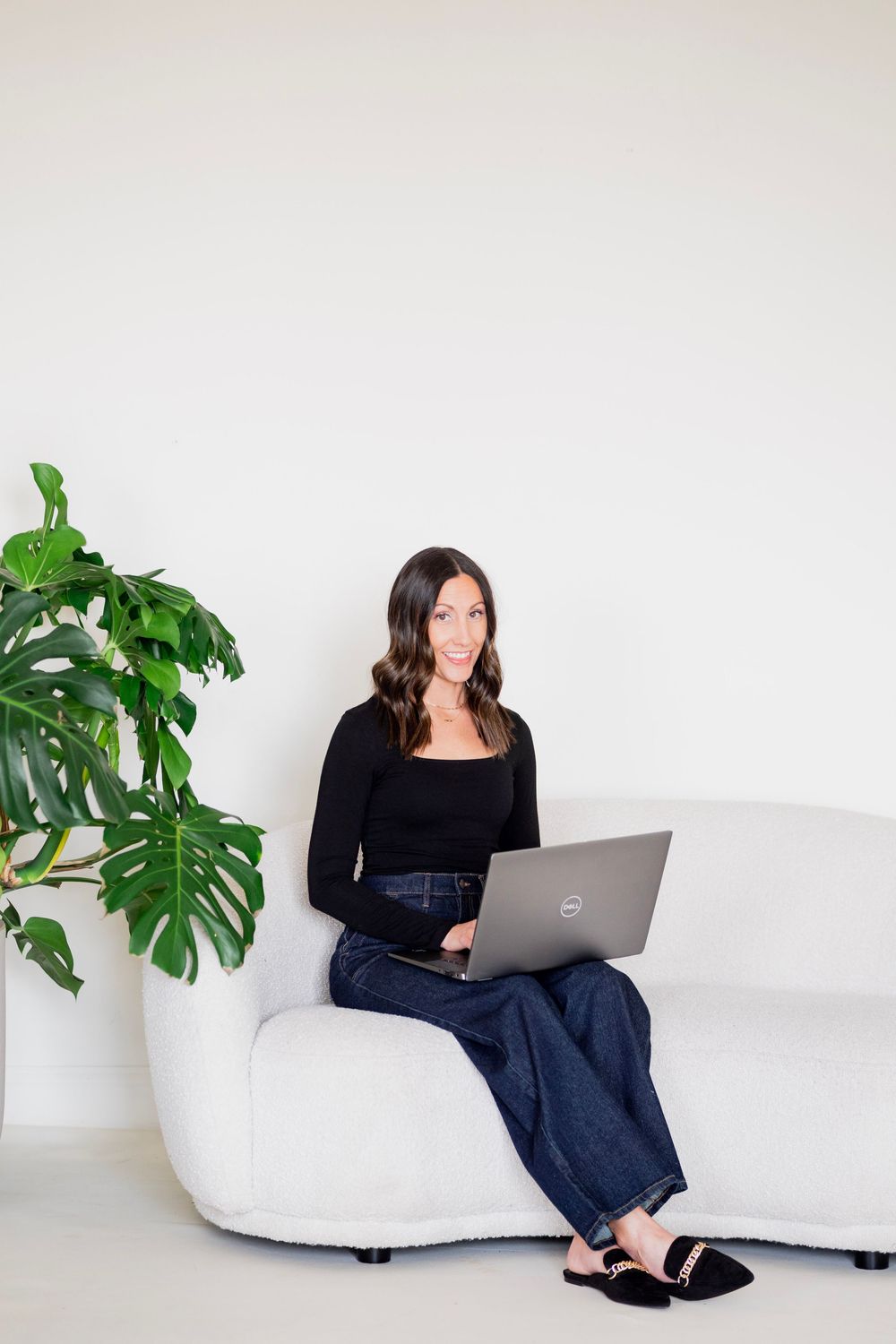 Brunette woman in black long-sleeve top and jeans sitting on white couch with Dell laptop.