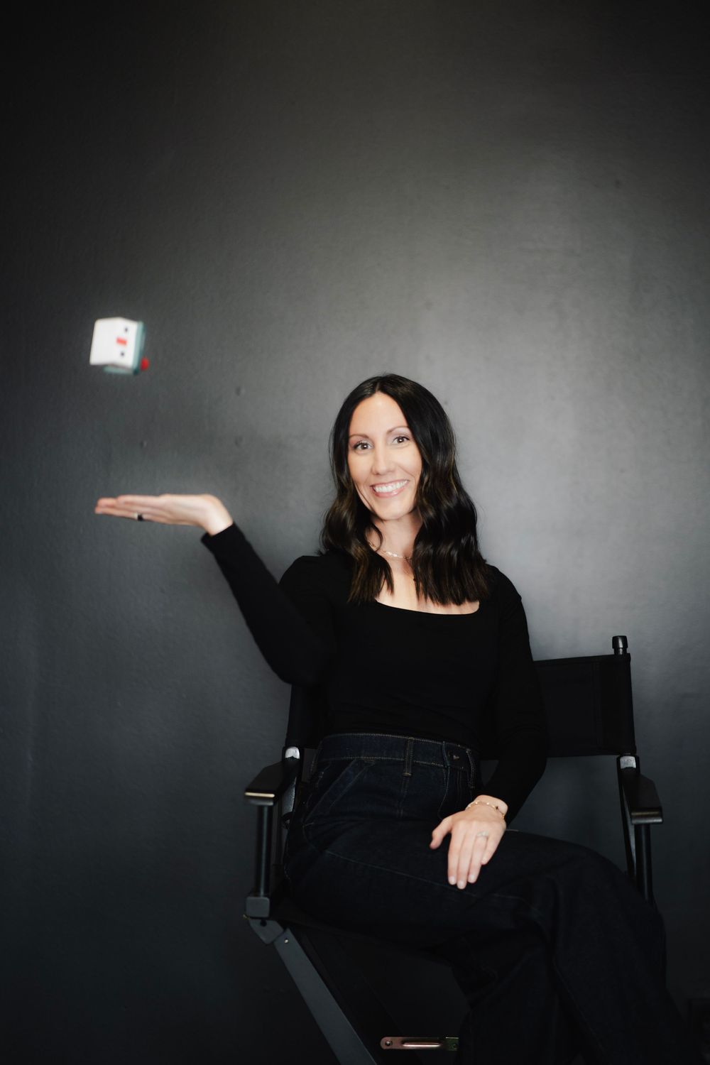 Woman in black outfit sitting in director's chair tossing a playing card in the air against dark background.