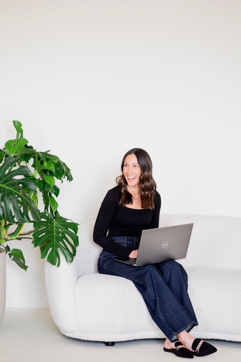 Smiling brunette woman in black top and jeans using Dell laptop on white couch near plant.
