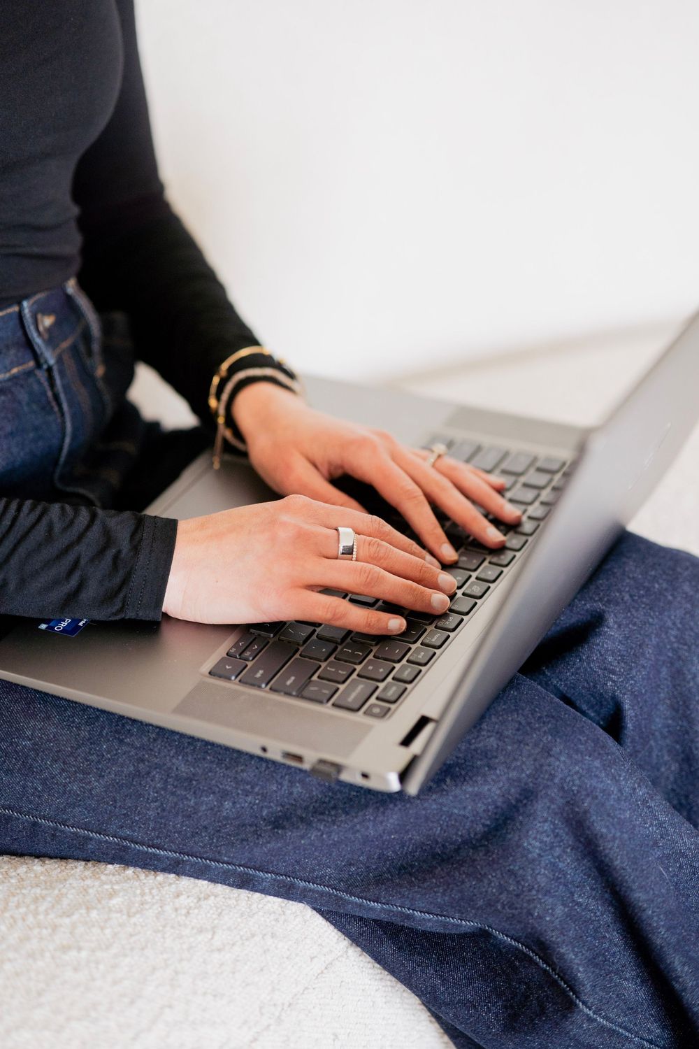 Close-up of woman's hands typing on a silver laptop while sitting on a white couch in jeans.