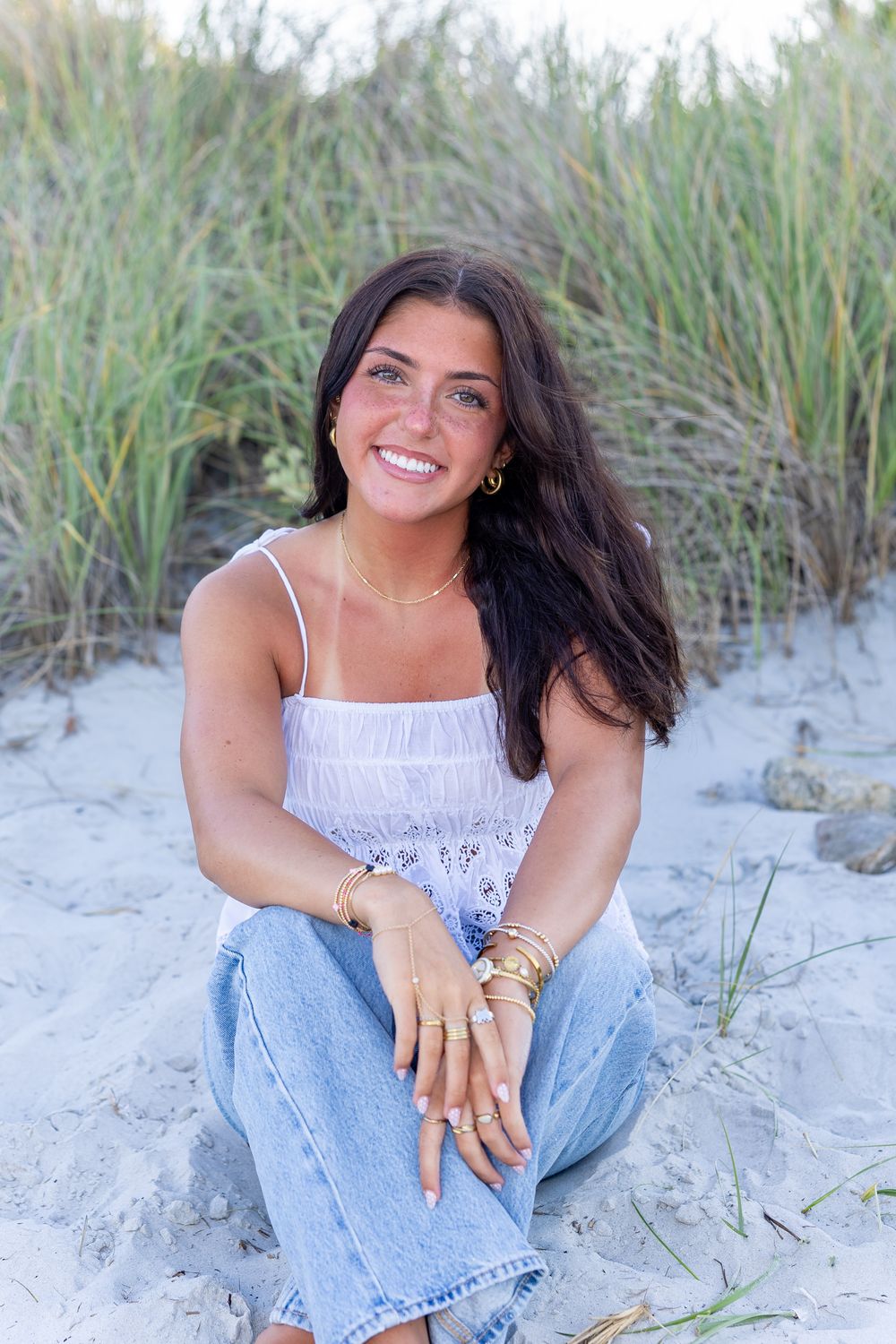 High school senior sits on sandy beach dunes, wearing white top, jeans, and gold jewelry, smiling brightly at Wingaersheek Beach- Gloucester
