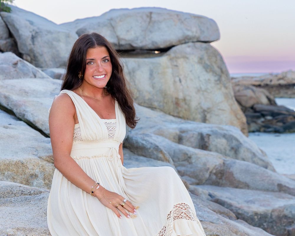 Woman in cream dress sitting on rocks at beach during poses at sunset during Senior photos on Wingaersheek Beach- Gloucester, MA.