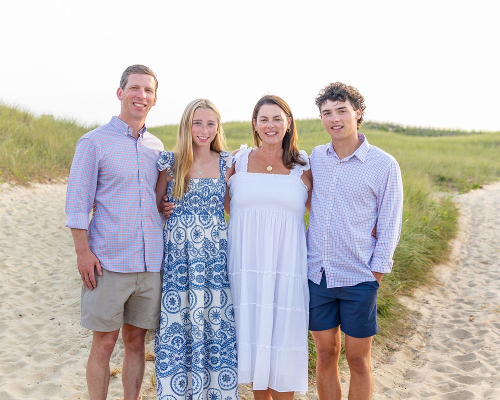 Family of four pose together on a sandy beach, dressed in coordinating blue & white outfits on a sunny day during family photos on Nantucket