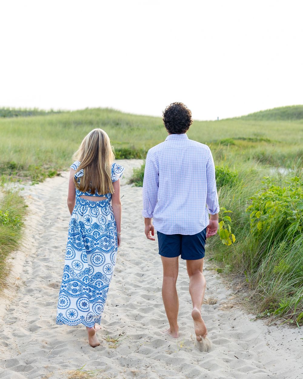 Siblings walk barefoot on sandy beach path surrounded green dunes in coordinated blue outfits during photos on Miacomet Beach Nantucket.