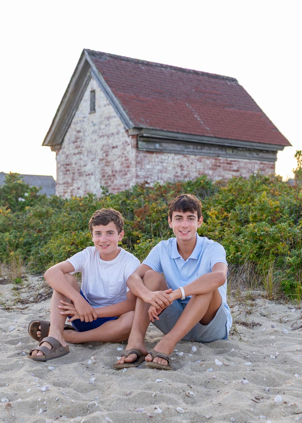 Two smiling boys sitting on beach by a rustic brick building with red roof during a family photo session at Brant Point Lighthouse Nantucket