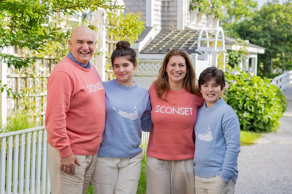 A family in matching blue & coral sweaters pose by coastal cottages & Sconset footbridge during a family portrait session on Nantucket, MA.