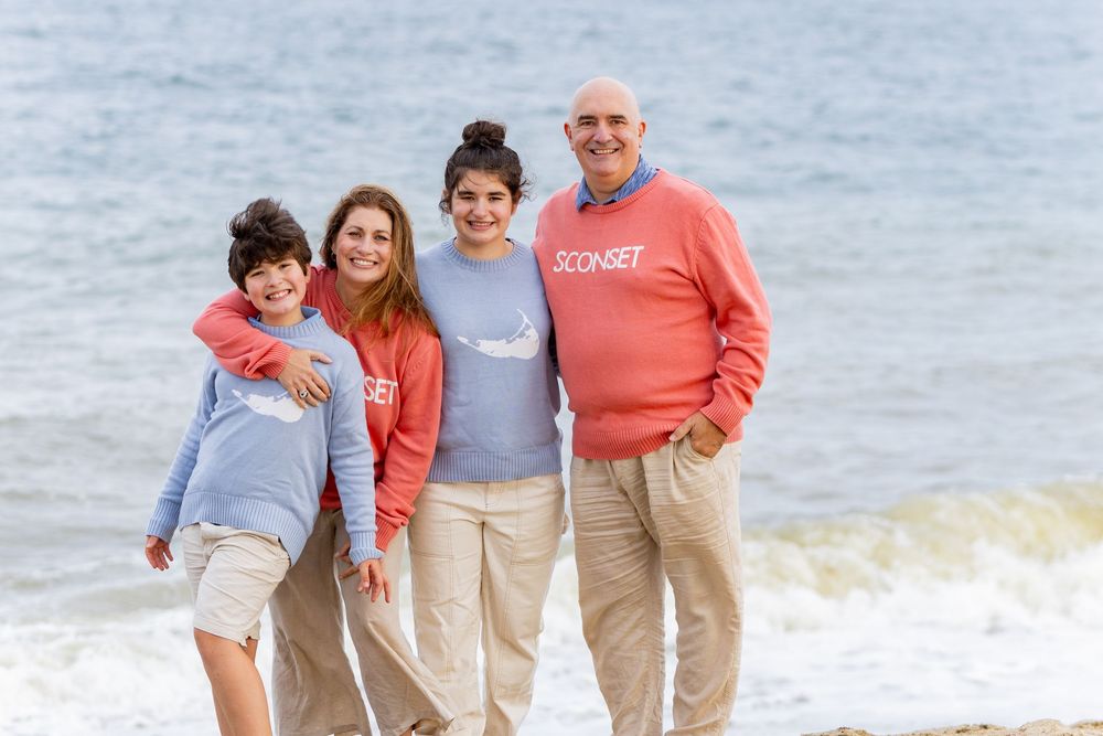 Family of four smiling on a beach, wearing coordinating blue and coral Sconset sweaters with ocean waves behind them on Nantucket, MA.