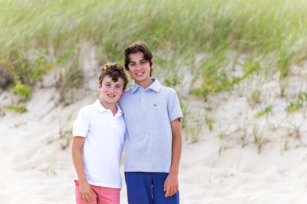 Siblings pose together on a sandy beach with grass-covered dunes in the background during family photos in Chatham, MA on Cape Cod.