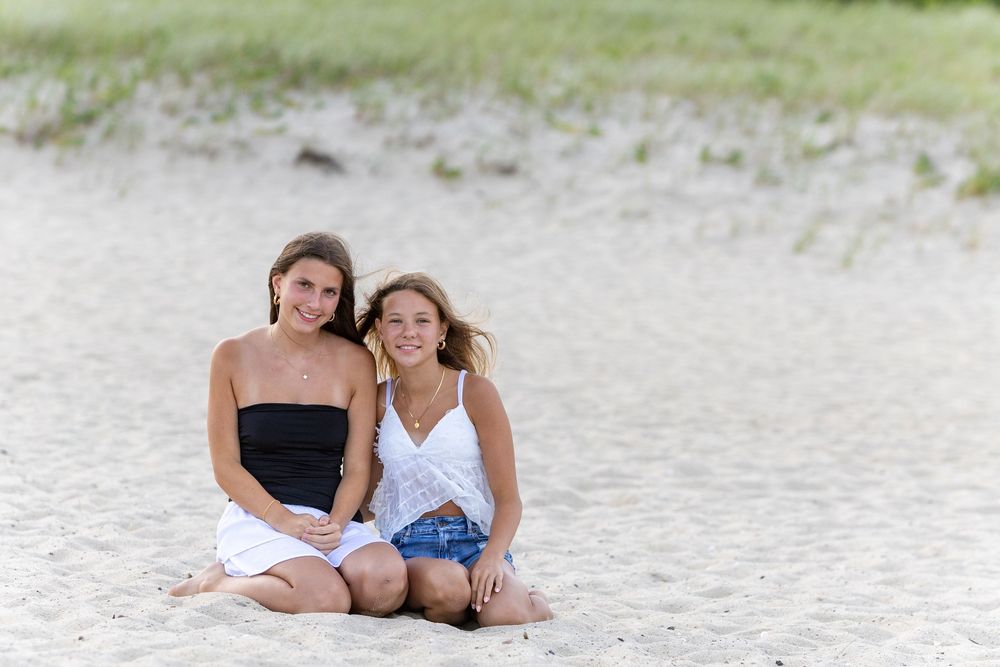 Friends sit together on a sandy beach with sea grass smiling, one in black top and one in white top on Lighthouse Beach in Chatham, MA.