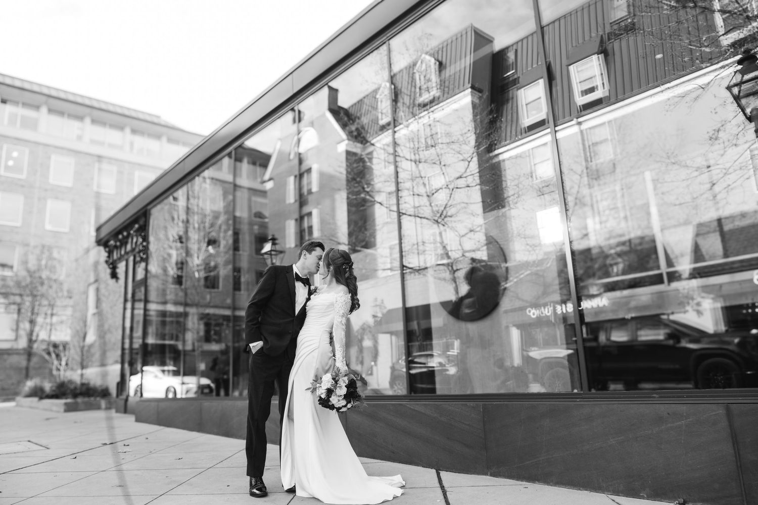 Black and white photo of bride and groom sharing a kiss outside modern glass building.