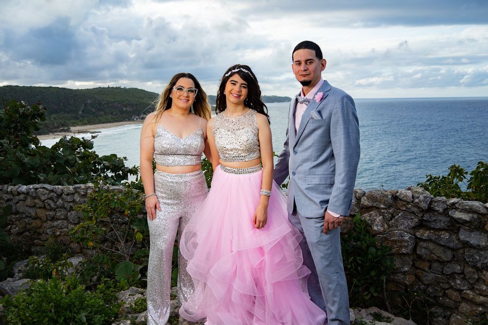 Three teens in formal attire pose outdoors with a scenic ocean and rocky coastline backdrop.