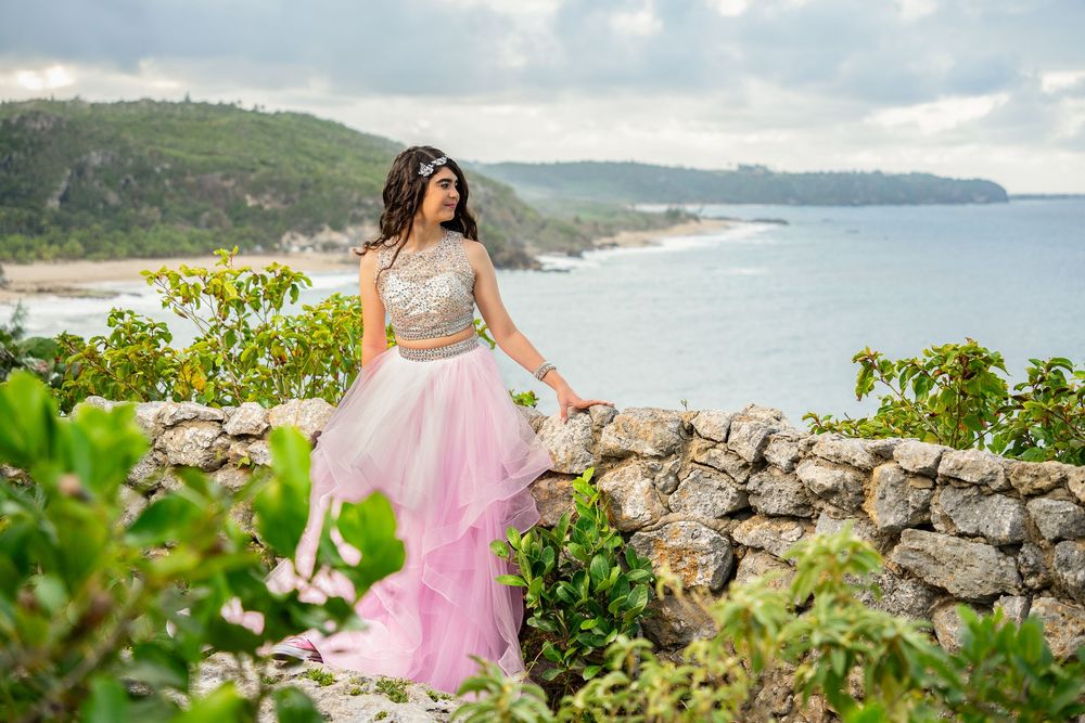Woman in pink tulle skirt and gold top poses by stone wall with ocean and green hills backdrop.