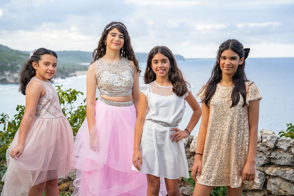 Four young girls in elegant dresses pose together outdoors with a scenic ocean and hillside view behind them.
