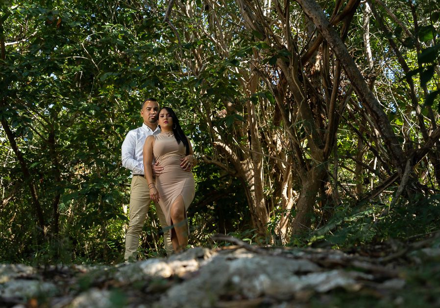 Couple embracing in a lush green forest, woman in beige dress, man in white shirt, romantic outdoor portrait.