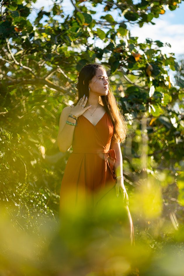 Woman in rust orange dress standing among lush green trees in golden sunlight, eyes closed peacefully.