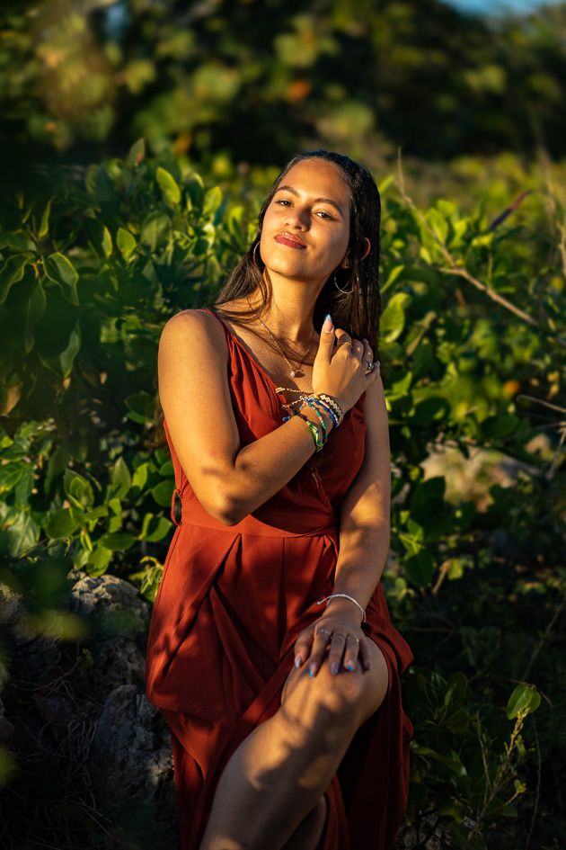Woman in red dress poses gracefully among lush green tropical foliage in warm golden sunset light.