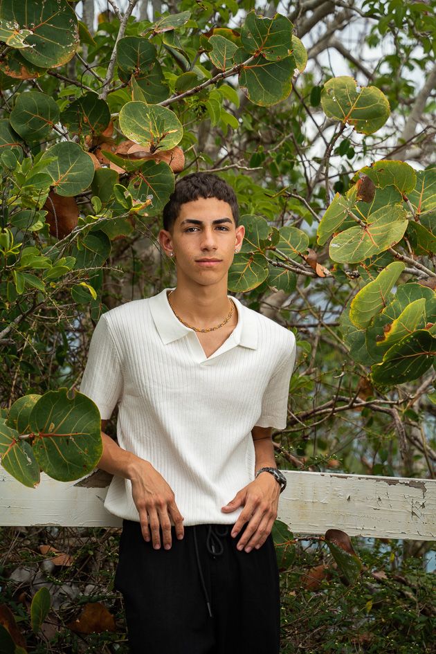 Young man in white ribbed polo shirt leans against white fence surrounded by lush green tropical foliage outdoors.