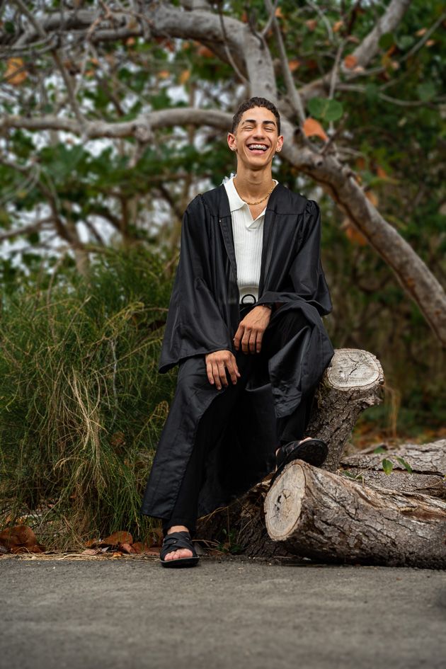 Young man in graduation gown smiling while sitting on a log outdoors with trees in the background.