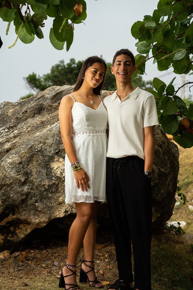 Young couple posing outdoors in semi-formal attire near a large rock and green foliage.