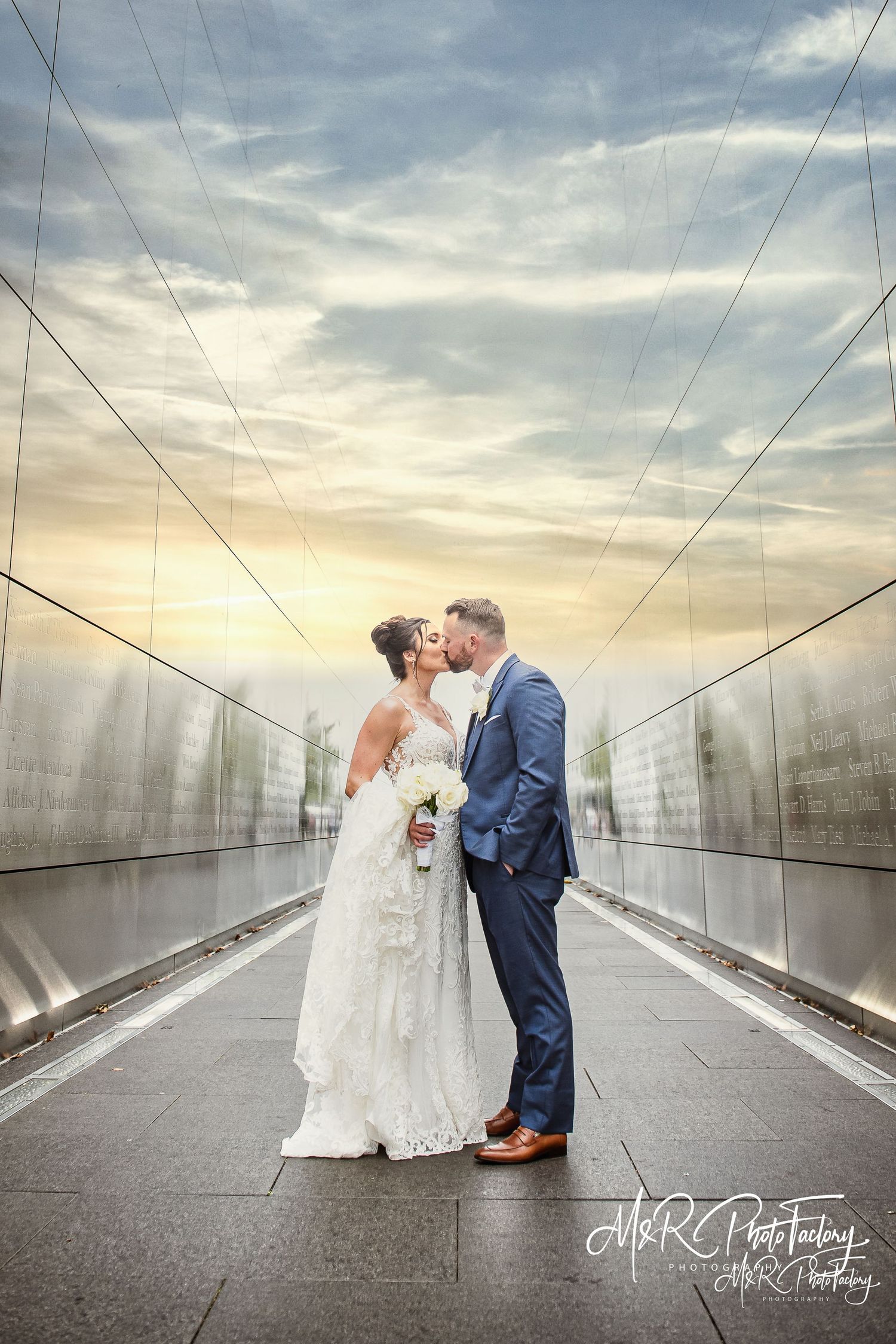 A bride and groom share a romantic kiss on a reflective walkway under a dramatic sunset sky.