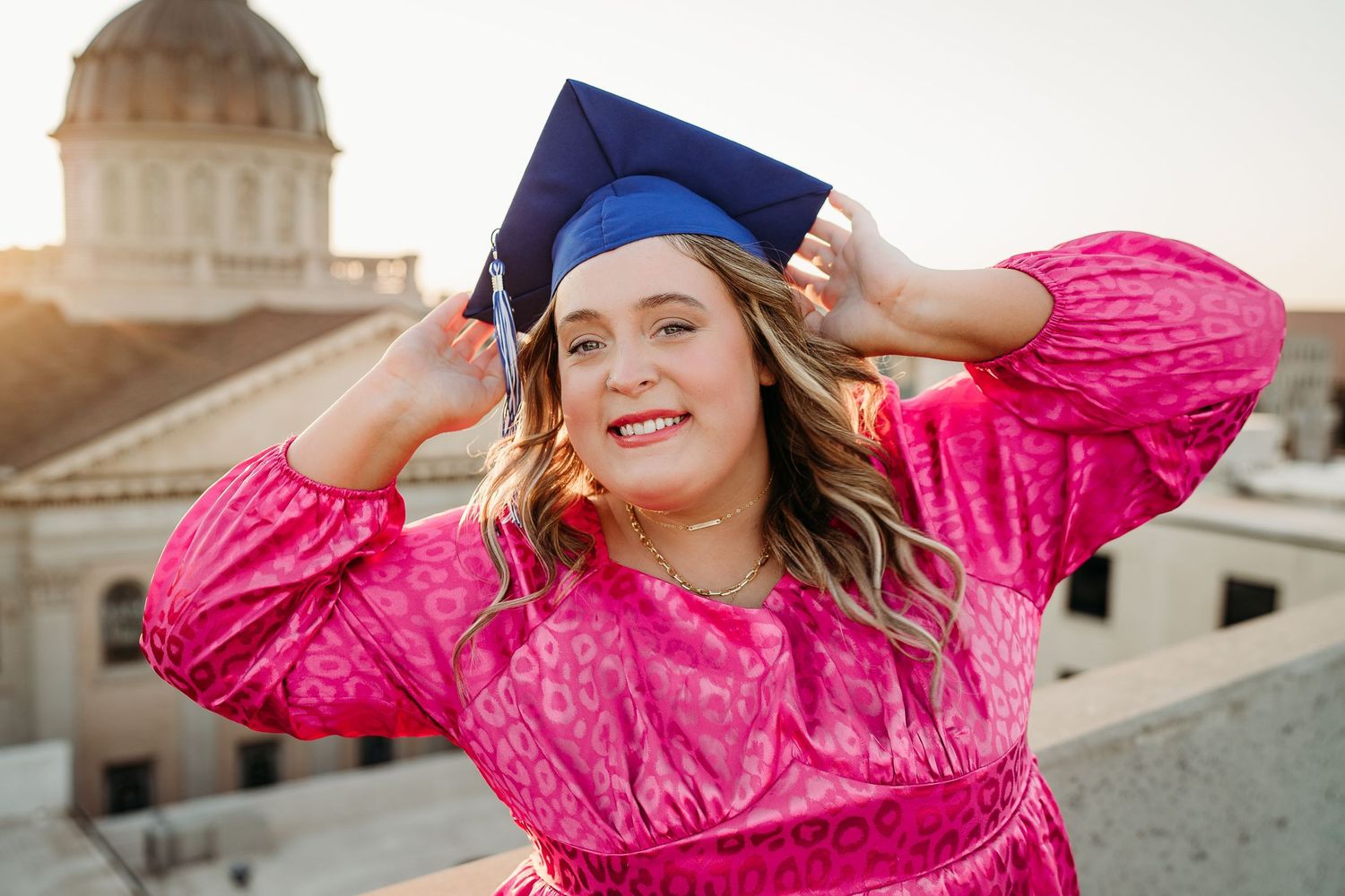 Young woman in pink dress adjusting blue graduation cap, smiling outdoors with golden hour sunlight and building dome behind her.
