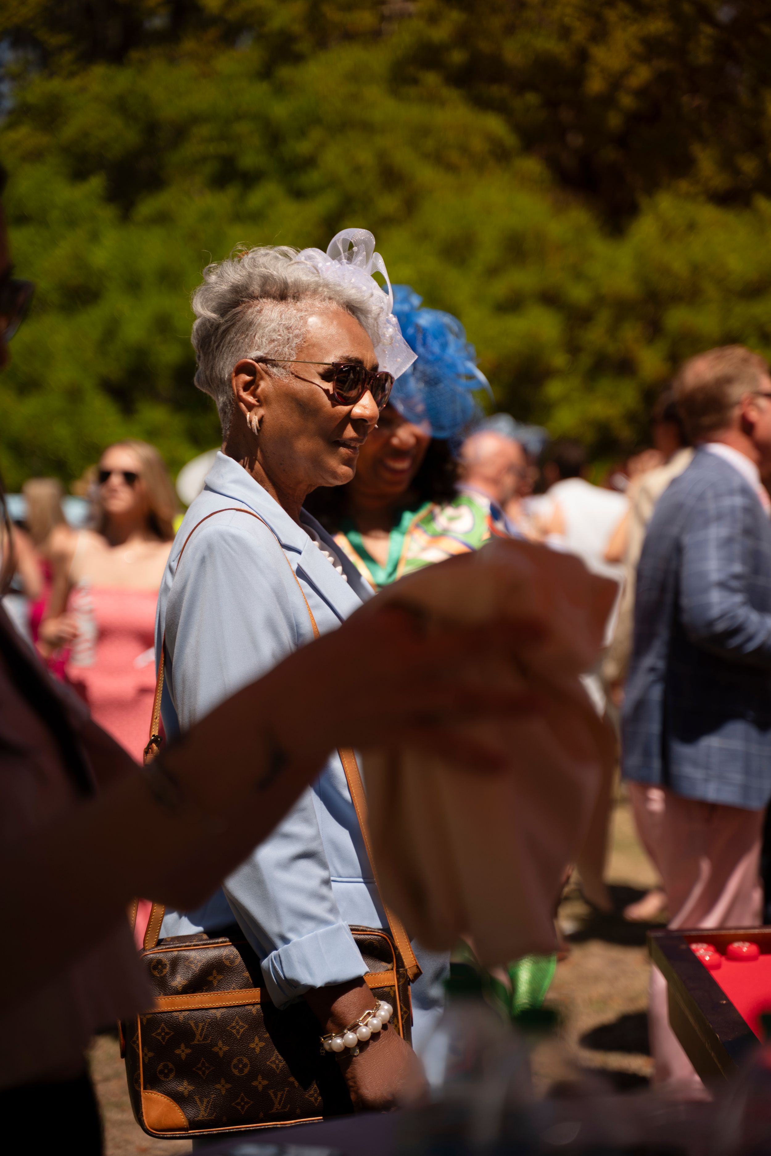 Elegant older woman with silver hair wearing sunglasses and blue jacket carrying Louis Vuitton bag at outdoor garden party.