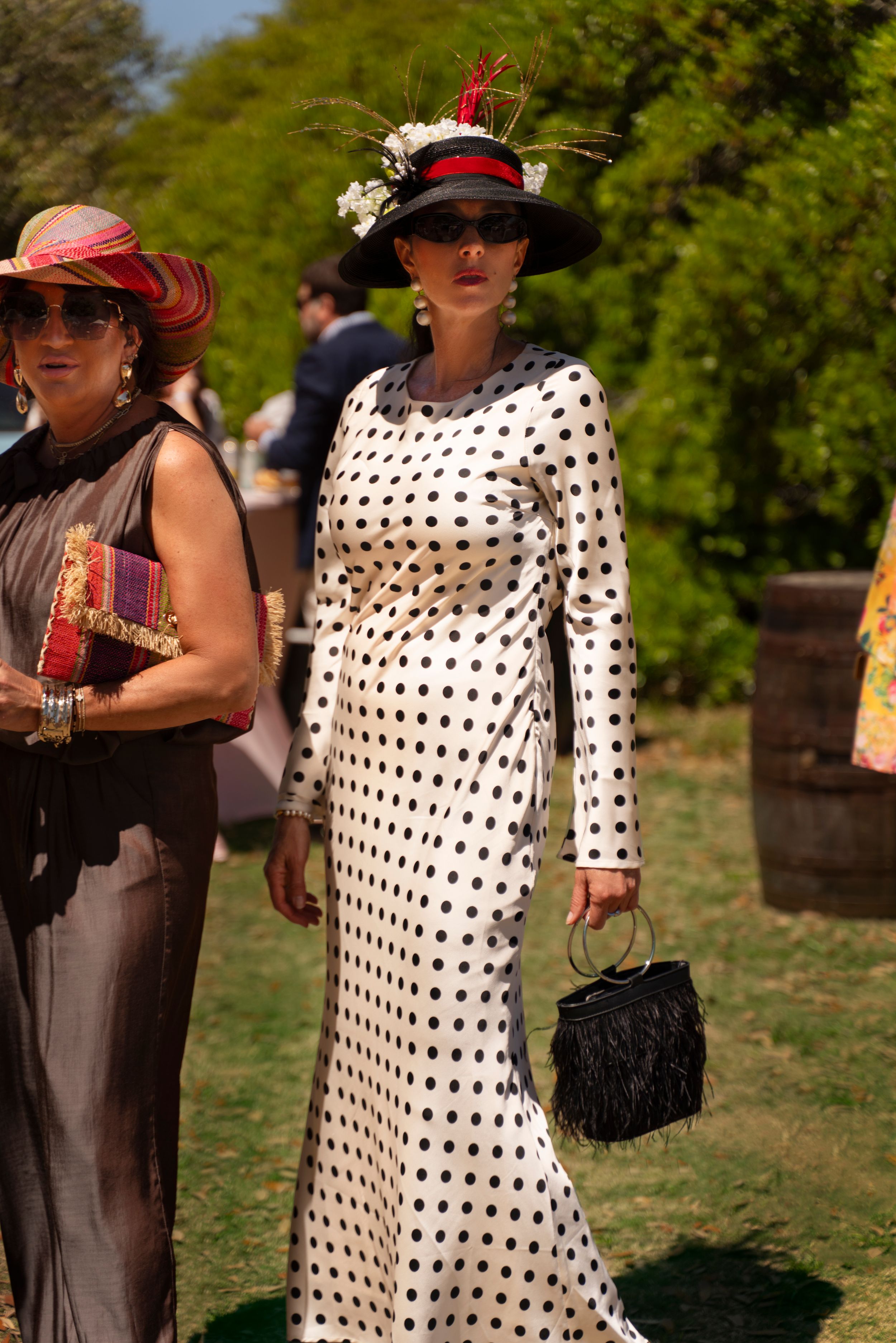 Woman in white polka dot dress and black wide-brim hat with floral detail carries black fringe bag at outdoor event.