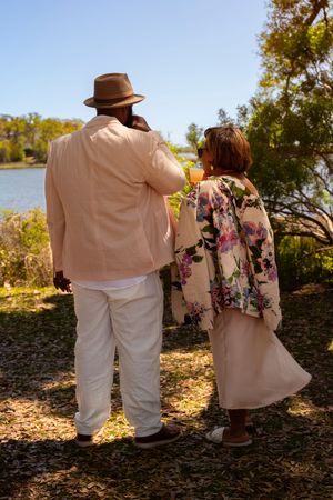 Couple in elegant summer attire stand by a sunlit lake, man in beige suit and hat, woman in floral jacket.